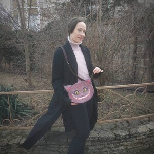 May include: A woman wearing a black coat and pink sweater holds a pink purse with a grinning cat face. The purse has a black strap and is worn over the shoulder.