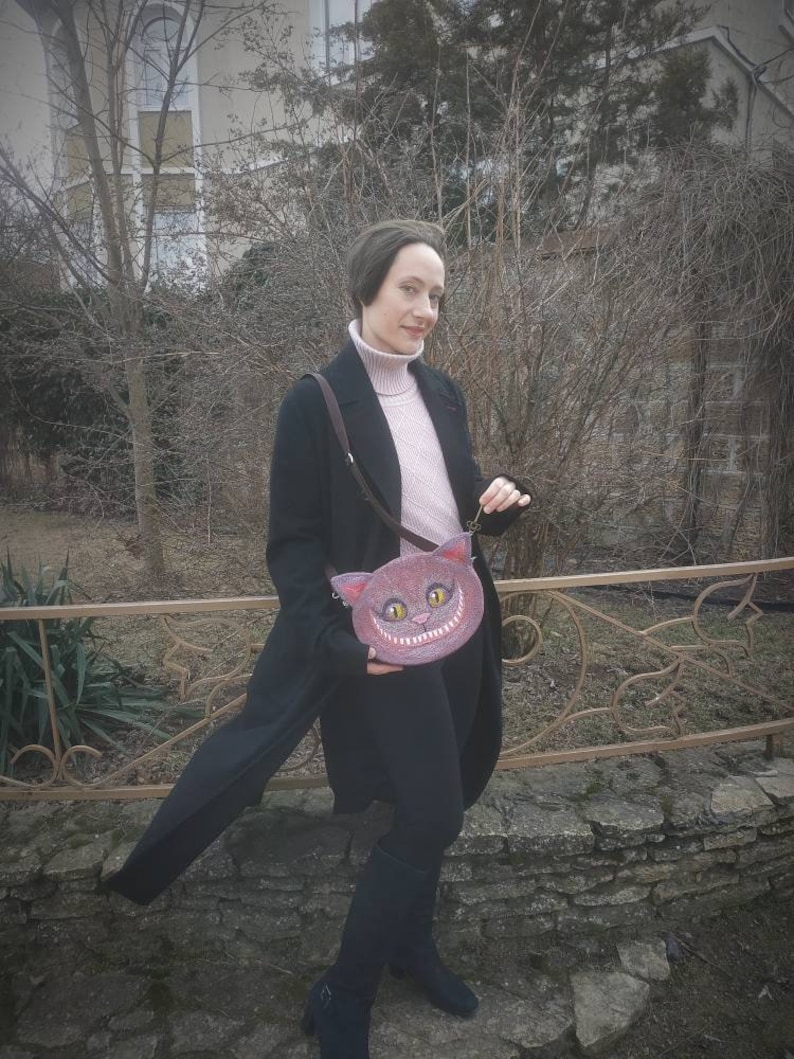 May include: A woman wearing a black coat and pink sweater holds a pink purse with a grinning cat face. The purse has a black strap and is worn over the shoulder.