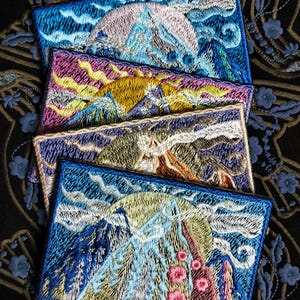 Può includere: Quattro toppe ricamate che rappresentano una scena di montagna in diverse palette di colori. Le toppe sono sovrapposte, con la toppa superiore che mostra una montagna blu con nuvole bianche e un cielo rosa. La seconda toppa mostra una montagna gialla con nuvole viola e un cielo rosa. La terza toppa mostra una montagna marrone con nuvole viola e un cielo blu. La toppa inferiore mostra una montagna blu con nuvole bianche e un cielo giallo.