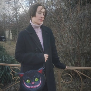 May include: A woman wearing a black coat and a pink turtleneck sweater is holding a black purse with a grinning Cheshire Cat design.