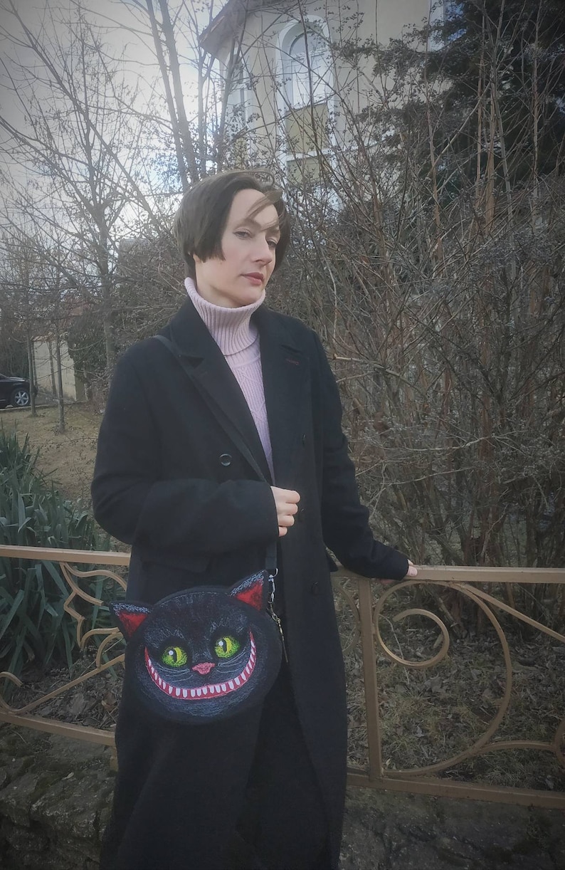 May include: A woman wearing a black coat and a pink turtleneck sweater is holding a black purse with a grinning Cheshire Cat design.