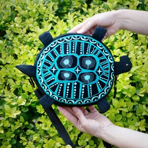 May include: A black and turquoise turtle-shaped crossbody bag with a geometric design. The bag has a black strap and a metal clasp.