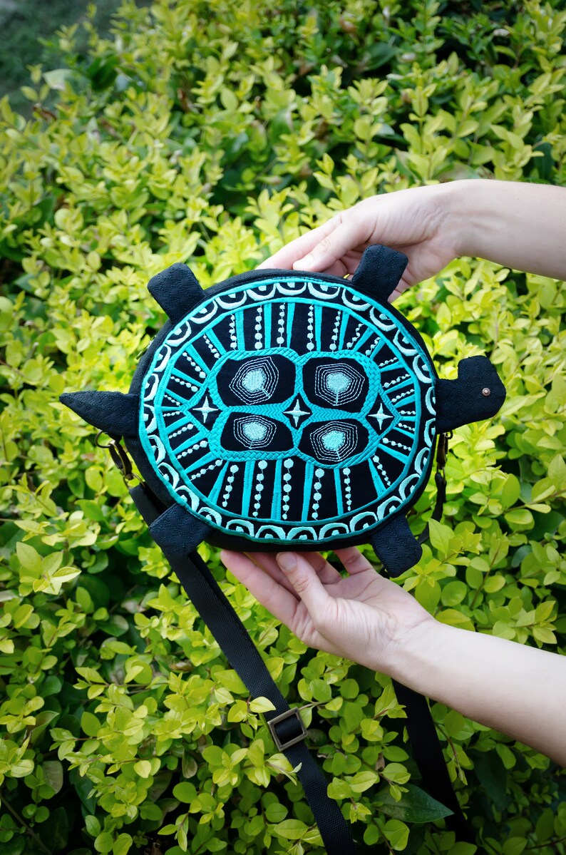 May include: A black and turquoise turtle-shaped crossbody bag with a geometric design. The bag has a black strap and a metal clasp.