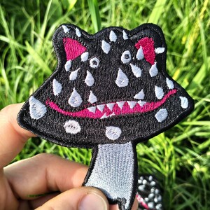 Può includere: Toppa a forma di fungo nero ricamata con un volto sorridente, punti bianchi e dettagli rosa. La toppa ha un gambo grigio ed è tenuta su uno sfondo di erba verde. La toppa è larga circa 7,6 cm.