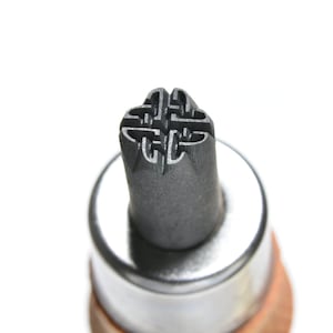 May include: Close-up of a metal stamp with a Celtic knot design. The stamp has a dark gray finish and is mounted on a wooden handle. The intricate knot pattern is clearly visible against the white background.