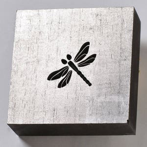 May include: A silver metal block with a black dragonfly design. The block has a textured surface.