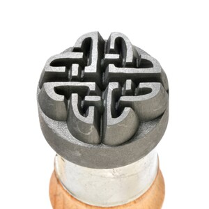 May include: A close-up of a metal stamp with a Celtic knot design. The stamp is mounted on a wooden handle.