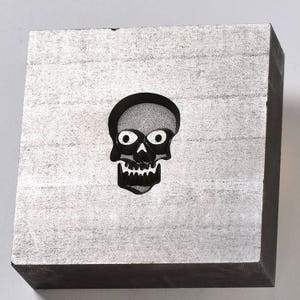 May include: A grey metal block with a black skull cutout. The skull has white eyes and a toothy grin.