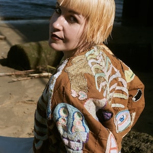 May include: A brown quilted jacket with colorful embroidered designs. The jacket features intricate patterns in shades of blue, green, gold, and pink. The jacket is worn by a person with short blonde hair.