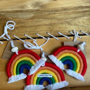 May include: Three handmade rainbow ornaments with red, orange, yellow, green, and blue arches. Each rainbow has a white fringe and a small tag. The text "Little Sunshine Rae" is on one of the rainbows. The ornaments are displayed on a wooden surface.
