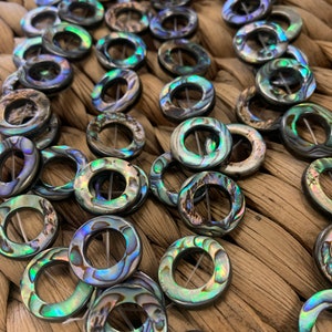 May include: A close-up of a strand of iridescent abalone shell beads. The beads are round with a hole in the center and have a dark green, blue, and purple color.