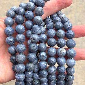 Op de afbeelding: Een ketting of armband van blauwgrijze kralen. De ronde kralen hebben een textuur en poreus oppervlak. De sieraden worden in een hand gehouden, waarbij de kleur en textuur worden benadrukt. De kralen zijn aan elkaar geregen.