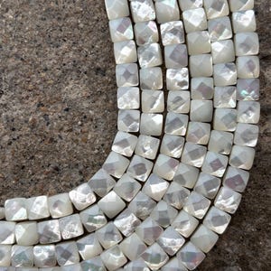 May include: Close-up of a strand of square, iridescent white beads. The beads have a faceted surface, catching the light and creating a shimmering effect. The beads are strung together, forming a curved shape against a textured, gray background.