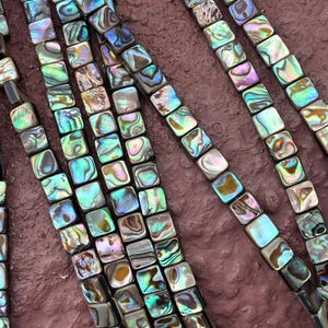 Puede incluir: Primer plano de varios hilos de cuentas cuadradas de concha de abulón. Las cuentas muestran colores iridiscentes de azul, verde y morado, con acentos marrones y negros. Las cuentas están ensartadas, creando un patrón texturizado y colorido.