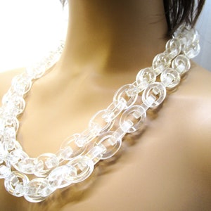 Può includere: Una collana di plastica trasparente con grandi perline circolari collegate. La collana è a strati, creando un aspetto spesso e massiccio.