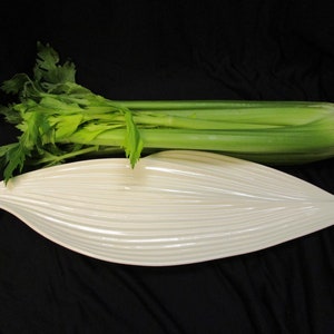 Może przedstawiać: Biała ceramiczna tacka w kształcie liścia z żebrowaną fakturą. Pęczek selera umieszczony jest na czarnym tle za tacką.
