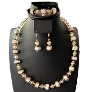 Puede incluir: Un conjunto de joyas que incluye un collar, una pulsera y pendientes. El collar y la pulsera están hechos de cuentas de vidrio transparente y marrón con detalles dorados. Los pendientes están hechos de cuentas de vidrio transparente con detalles dorados.