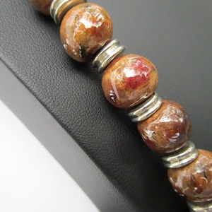 Puede incluir: Un brazalete de cuentas de madera marrón con separadores de metal plateado. Las cuentas están pintadas con diseños florales blancos y rojos.