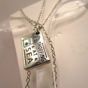 Op de afbeelding: Zilveren ketting met een bedel waarop "Shopping at Sea" staat met een klein blauw steentje. De bedel is gevormd als een ticket of een label.