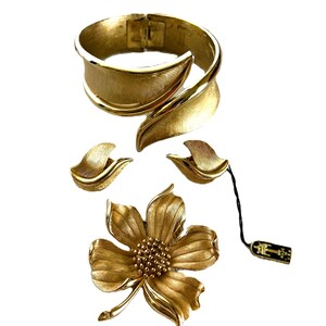 May include: A gold-toned jewelry set featuring a flower brooch, a pair of leaf-shaped earrings, and a wide, curved bangle bracelet.
