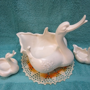 May include: Three white ceramic swan-shaped planters. The largest planter is a bowl with a wide opening and a swan's head and neck forming the rim. The two smaller planters are more compact and have a more traditional swan shape. The planters are sitting on a white crocheted doily.