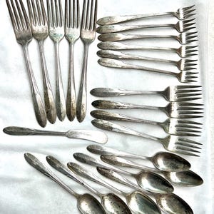 May include: An assortment of vintage silverware, including forks, spoons, and butter knives, arranged on a white surface. The silverware has a silver finish and shows signs of age and use, suggesting a classic or antique style.