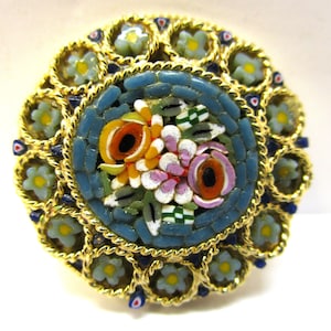Op de afbeelding: Een ronde, goudkleurige broche met een mozaïekontwerp met een cluster van kleurrijke bloemen. De bloemen zijn gemaakt van kleine glazen stukjes in tinten oranje, geel, roze en wit. De broche is omgeven door een decoratieve rand van goudkleurig metaal en kleine witte bloemen.