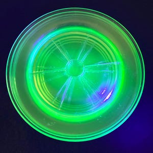 May include: A round, clear glass plate glows bright green under a black light. The plate has a circular design in the center with radiating lines. The rim of the plate is also a bright green color. The background is a dark blue.