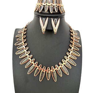 Puede incluir: Un conjunto de collar, pulsera y pendientes en tono dorado con un diseño geométrico. Las piezas de joyería están hechas de metal con un acabado texturizado.