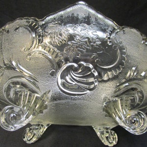 Könnte beinhalten: Eine klare Glasschüssel mit einem dekorativen, geätzten Design und drei Beinen. Die Schüssel hat einen gewellten Rand und ein Blumenmuster.