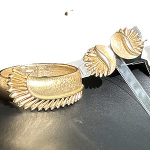 May include: A gold-toned jewelry set featuring a cuff bracelet and matching clip-on earrings. The bracelet has a textured surface with a sunburst design on one side. The earrings have a similar design, with a textured, fan-like pattern.