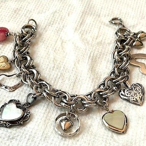 May include: A silver charm bracelet featuring a variety of heart-shaped charms, including a pink heart, a gold heart, a white heart, and a silver heart with a clover design.