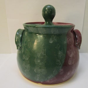May include: A green and burgundy ceramic crock with a lid and two handles. The crock has a speckled glaze.
