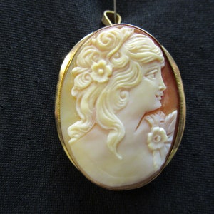 Puede incluir: Un colgante de camafeo ovalado dorado con un camafeo de concha tallada que representa el perfil de una mujer con una flor en el pelo.