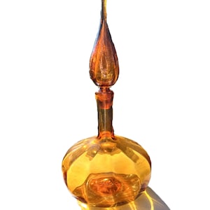 Pode incluir: Garrafa de vidro âmbar com base bulbosa e rolha longa em forma de gota. A garrafa tem uma superfície texturizada e um design decorativo. O vidro reflete a luz, criando sombras. A garrafa é provavelmente para fins decorativos.