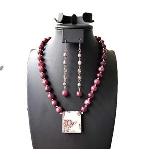 Puede incluir: Un collar de cuentas color burdeos con un colgante cuadrado con un diseño floral. Pendientes a juego con cuentas plateadas y cristales transparentes que cuelgan de ganchos plateados.