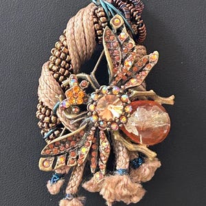 Puede incluir: Colgante artesanal con un gran cristal central facetado rodeado de cristales más pequeños y adornos metálicos. El colgante incorpora cordón trenzado color beige, cuentas color cobre y una piedra ámbar en forma de lágrima. El diseño incluye detalles en azul y marrón.