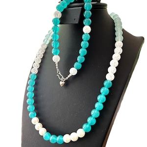 Puede incluir: Collar de cuentas redondas turquesas y blancas, exhibido en un maniquí negro. El collar tiene un cierre plateado con un dije en forma de corazón. Las cuentas hacen una transición de transparente a turquesa. Una joya.