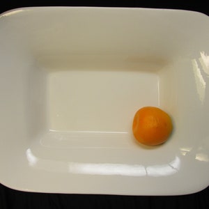Puede incluir: Un plato rectangular de cerámica blanca con una sola fruta naranja en el centro.