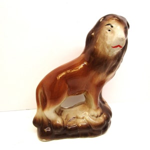 May include: A brown ceramic lion figurine with a white face and red painted lips. The lion is standing on a brown base.