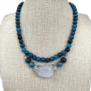 Puede incluir: Un collar de doble cadena con cuentas de piedras preciosas azules y un gran colgante de cuarzo transparente. El colgante está engastado en un metal plateado con un diseño ornamentado.