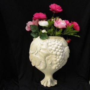 Puede incluir: Un jarrón de cerámica blanco con un diseño de vid y una base de pedestal. El jarrón está lleno de flores rosas y blancas.