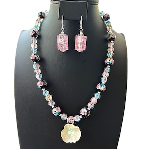Puede incluir: Un conjunto de collar y pendientes con un colgante floral y cuentas de colores. Los pendientes son cristales rectangulares rosas. Las cuentas son en tonos rosa, azul y negro. El conjunto se exhibe en un soporte negro.