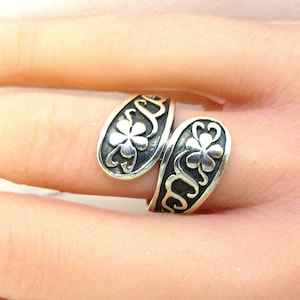 May include: A silver ring with a twisted design featuring two four-leaf clover motifs. The ring is worn on a finger.