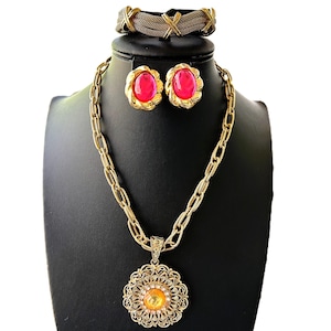 May include: A gold chain necklace with a large, round pendant featuring a yellow gem surrounded by a filigree design. The necklace is paired with a matching gold bracelet and a pair of pink gemstone earrings.