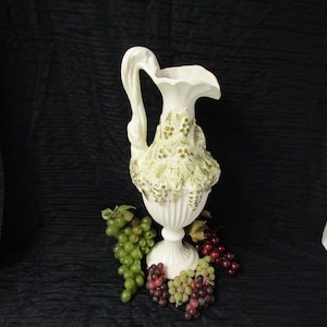 May include: A white ceramic pitcher with a gold and green grape design. The pitcher has a wide mouth and a handle. It is sitting on a black surface with green and red grapes.