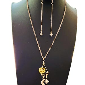 Puede incluir: Un conjunto de collar y pendientes dorados. El collar presenta una cadena con un colgante de una flor, una luna creciente y una estrella. Los pendientes son cadenas largas y colgantes con dijes en forma de estrella. La joyería se exhibe en un maniquí negro.