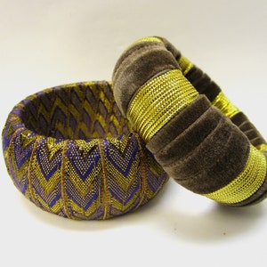 Op de afbeelding: Twee stoffen armbanden, een bruin met gouden rand en een met paars-gouden chevron-patroon.
