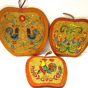 Puede incluir: Tres posavasos de madera con forma de manzana con diseños pintados. Los posavasos presentan un gallo, un diseño floral y un gallo con un corazón. Los posavasos están pintados en colores brillantes, incluyendo rojo, amarillo, azul y verde.