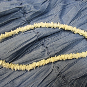 以下が含まれることがあります： 金色の留め具が付いた白い貝殻のネックレス。ネックレスは、小さな平らな貝殻のピースを繋げて作られています。